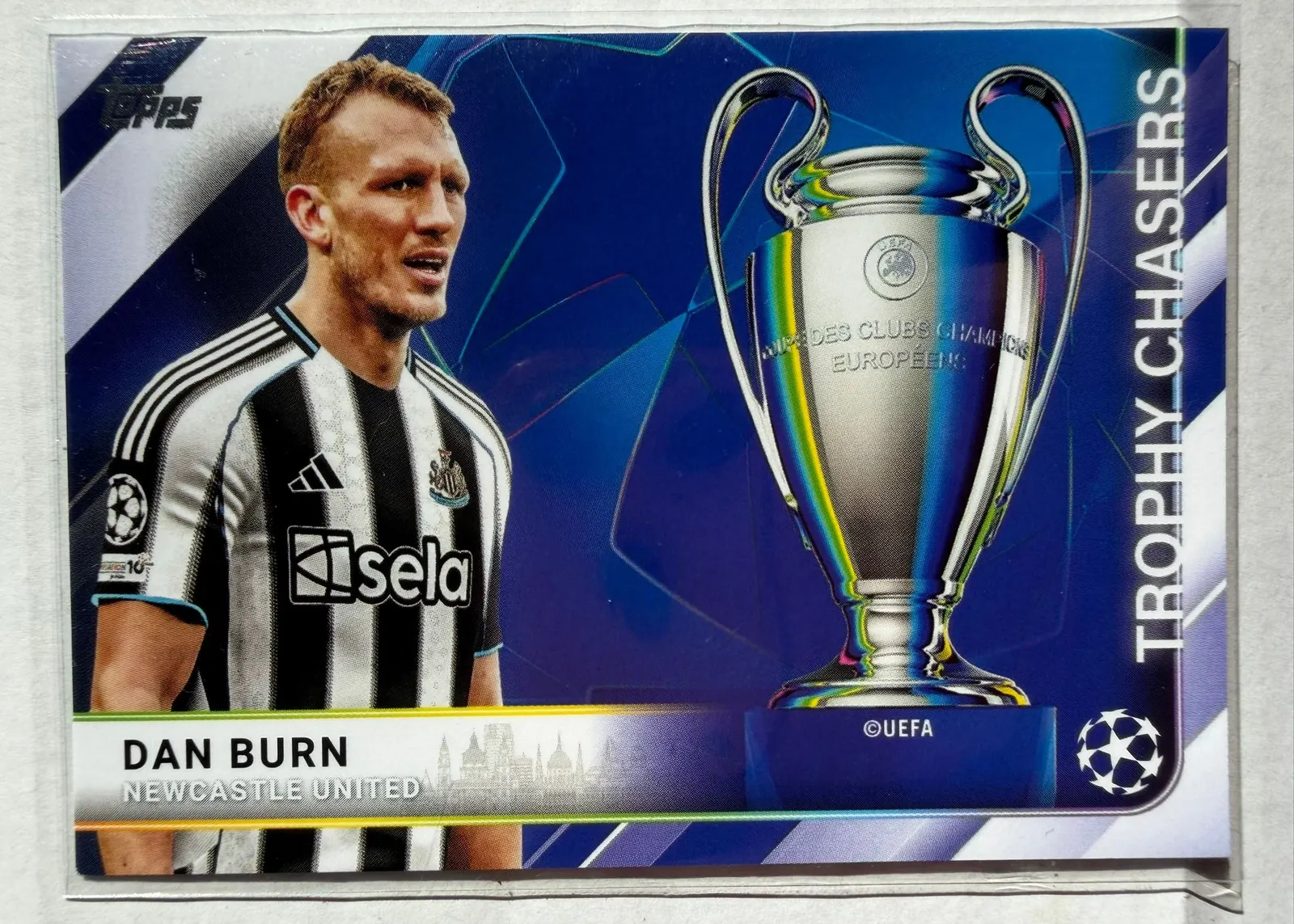 Dan Burn, Topps UEFA Club Competitions 2025/26 Trophy Chasers, karta TC-3 — awers poziomy z zawodnikiem w biało-czarnym stroju Newcastle United po lewej i pucharem Ligi Mistrzów po prawej
