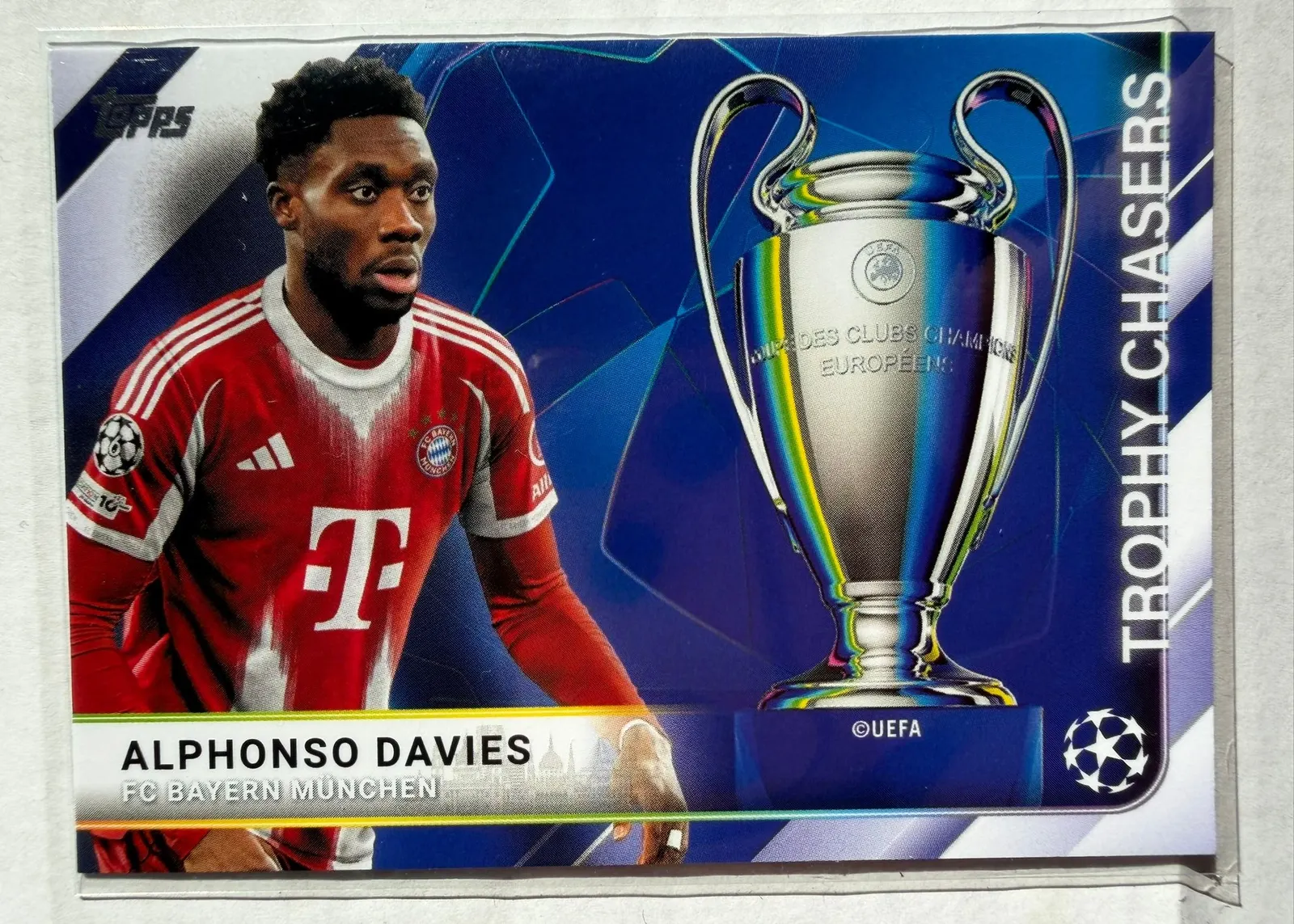 Alphonso Davies, Topps UEFA Club Competitions 2025/26 Trophy Chasers, karta TC-13 — awers poziomy z zawodnikiem w czerwonym stroju Bayernu po lewej i pucharem Ligi Mistrzów po prawej
