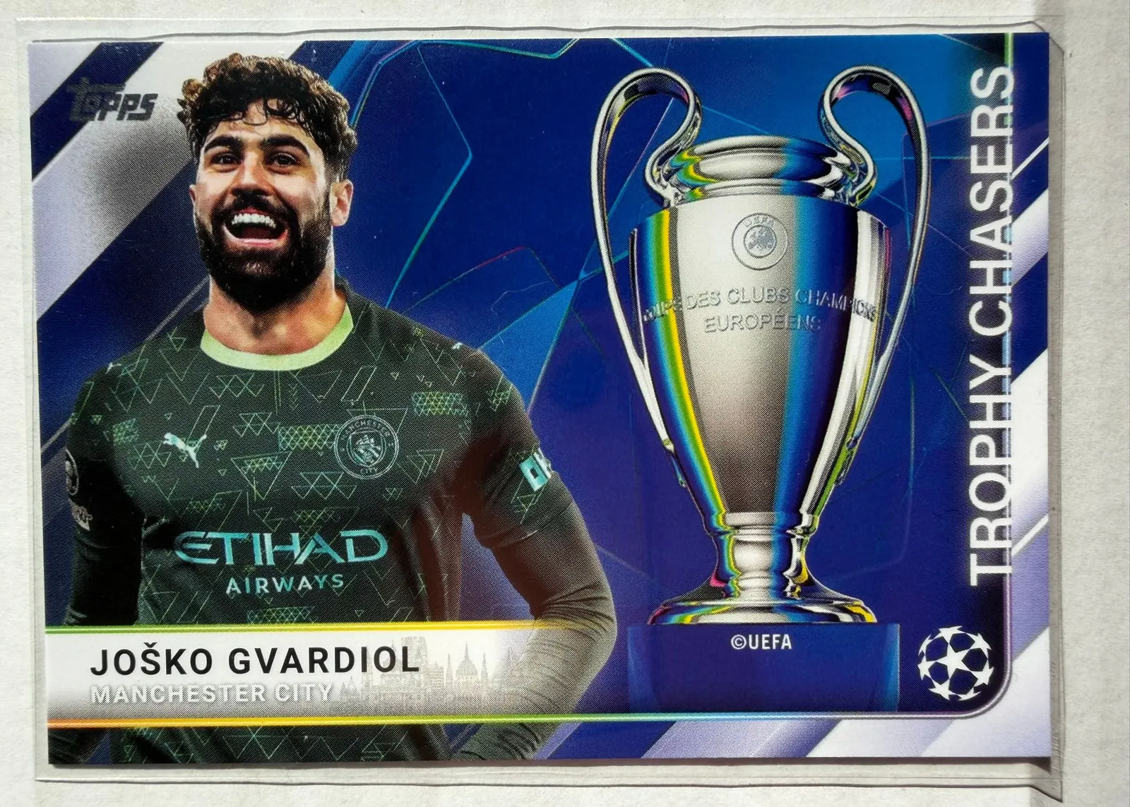 Joško Gvardiol, Topps UEFA Club Competitions 2025/26 Trophy Chasers, karta TC-4 — awers poziomy z zawodnikiem w ciemnym stroju Manchesteru City po lewej i pucharem Ligi Mistrzów po prawej